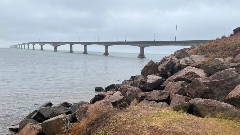 A large concrete bridge is seen in the Northumberland Straight, with red clay, green grass and large boulders in the foreground. 