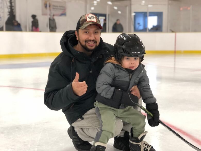 A man holds a child on his knee on a hockey arena and gives the camera a thumbs up. The child is wearing a hockey helmet and skates. 