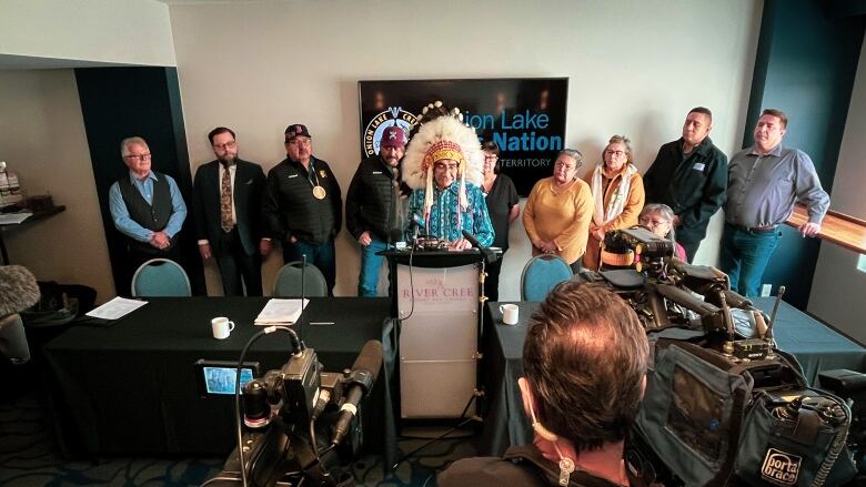 Onion Lake Cree Nation Chief Harry Lewis stands on a podium addressing the media. Council members and lawyers stand behind him.