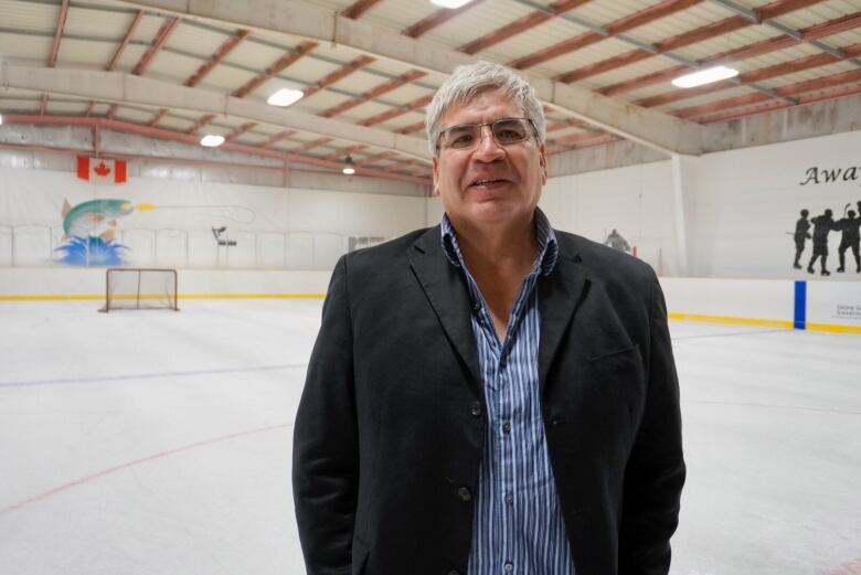 A man stands on an ice rink looking at the camera. The man is wearing a button up shirt and jacket.