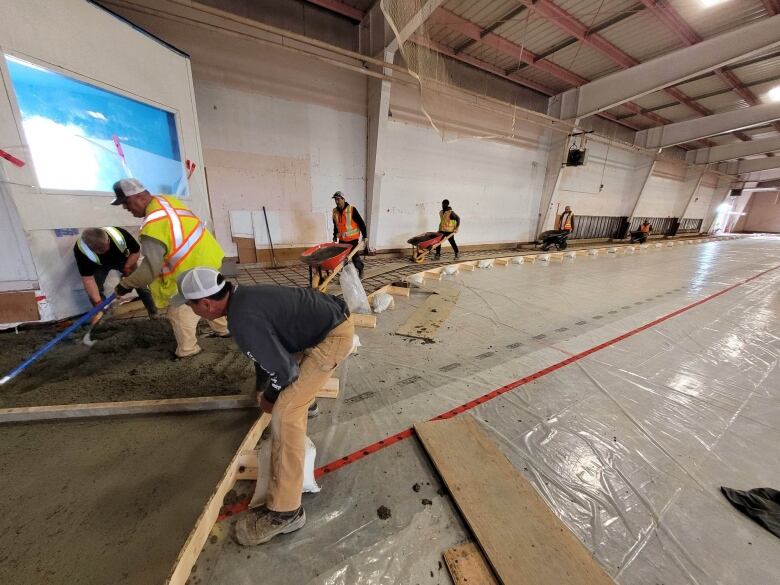 A construction crew pushes wheelbarrows of materials from a far side of an area into an area where the concrete is being smoothed across a surface.