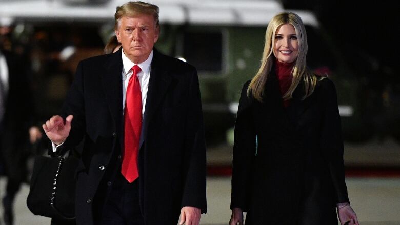 A man raises his right hand while walking besides a younger woman on the tarmac.