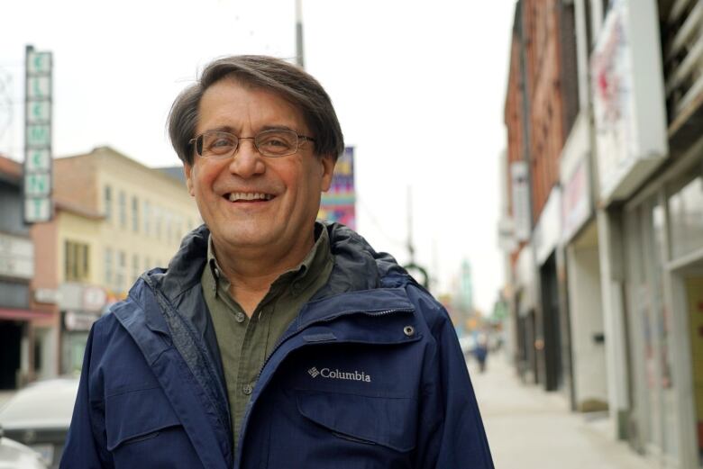 A man with dark hair and glasses stands outside wearing a blue jacket.