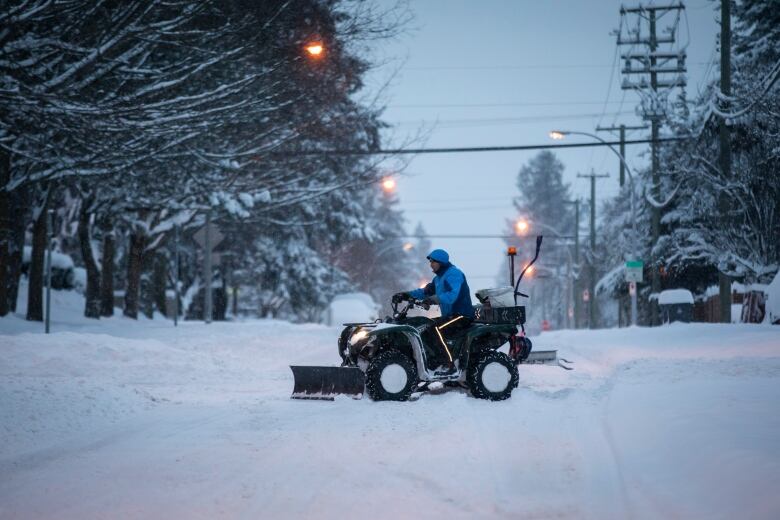 A person sits on an all-terrain vehicle equipped with a snow plow in a snowy, treed neighbourhood, helping to clear a snowbound street.