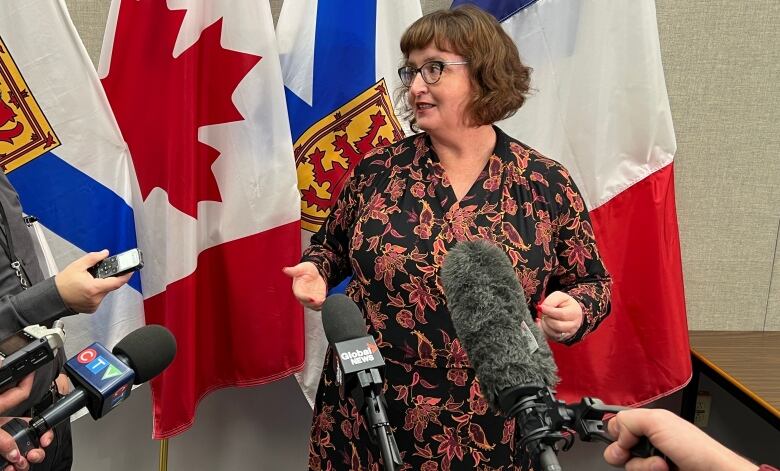 A politician with brown hair and glasses speaks to reporters who gather around with microphones.