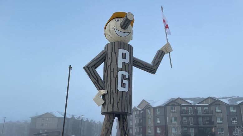 A wooden statue with the word 'PG' on its body stands in an open space with snow holding a flag.