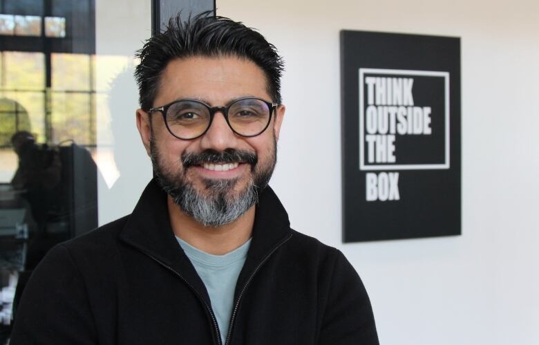 A man in black plastic-rimmed glasses faces the camera, next to a sign that says 