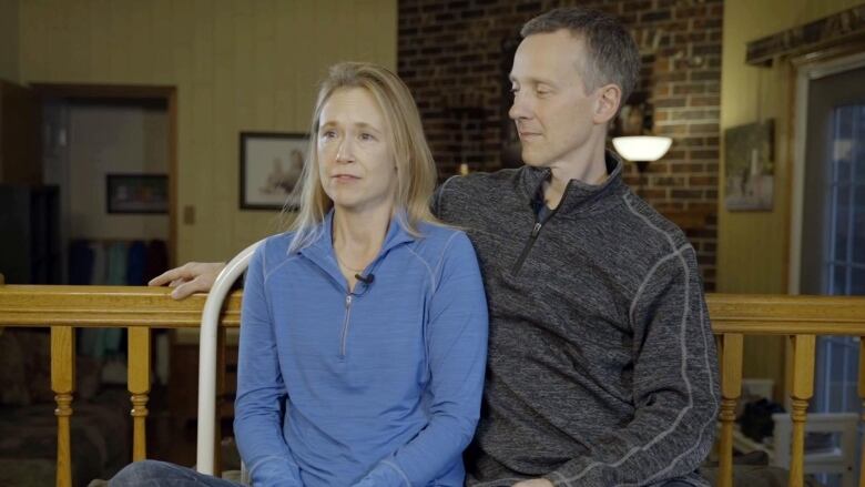 A seated woman looks to the left of the camera, and a man sits looking at her with his arm stretched out behind her.