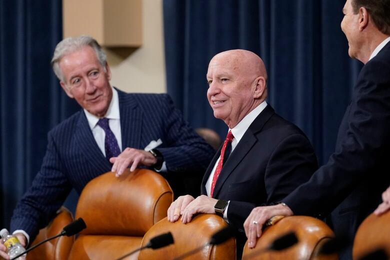 Two individuals wearing suits, one bald, one grey-haired, speaking to each other while standing behind leather chairs at a table. 