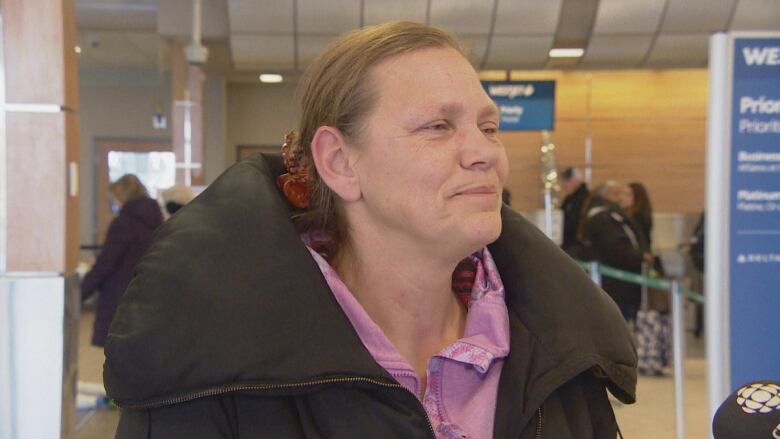 A woman with red hair is wearing a pink polo shirt with a brown coat. She is standing in an airport lobby.