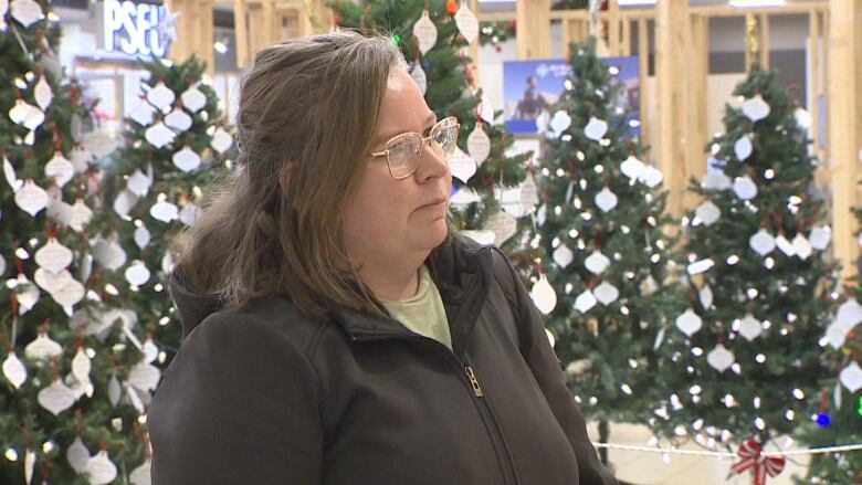 Woman with shoulder length brown hair wearing fleece jacket stands in mall in front of Christmas trees.