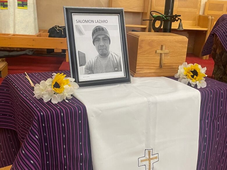 A black and white photo of Salomon Lzaro seen in the foreground. Beside it to the right is a wooden box with a cross containing his ashes. Black velvet bags can be seen in the background on the other side. They contain the ashes of other undocumented workers who also died of COVID-19