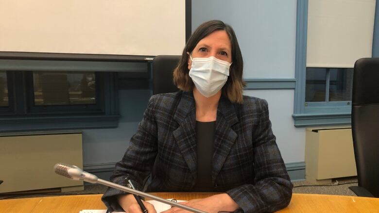 Dr. Vera Etches sits at a desk wearing a blue mask, a blue plaid jacket looking at the camera and holding a pen poised over a notebook. 