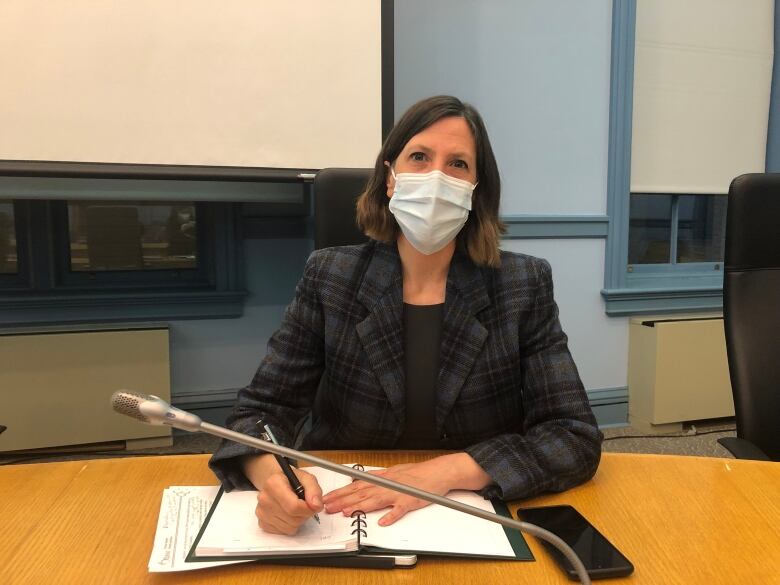 Dr. Vera Etches sits at a desk wearing a blue mask, a blue plaid jacket looking at the camera and holding a pen poised over a notebook. 