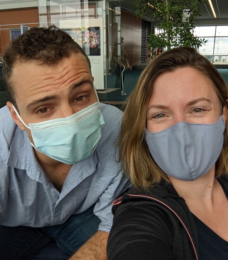 A couple wearing face maskes smile at the camera. One of them, a woman, is siting on a chair while a man leans over the chair beside her. 