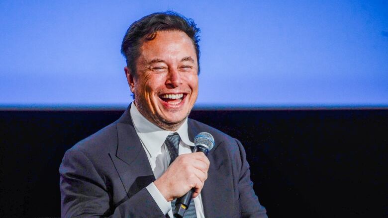 A seated man in a suit laughs while holding a microphone.