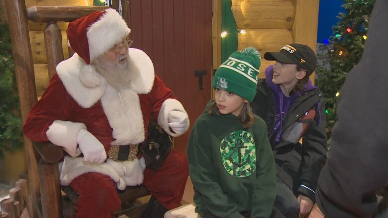 A man dressed up as Santa Claus sits in a department store talking to two children sitting in front of him for a picture. 