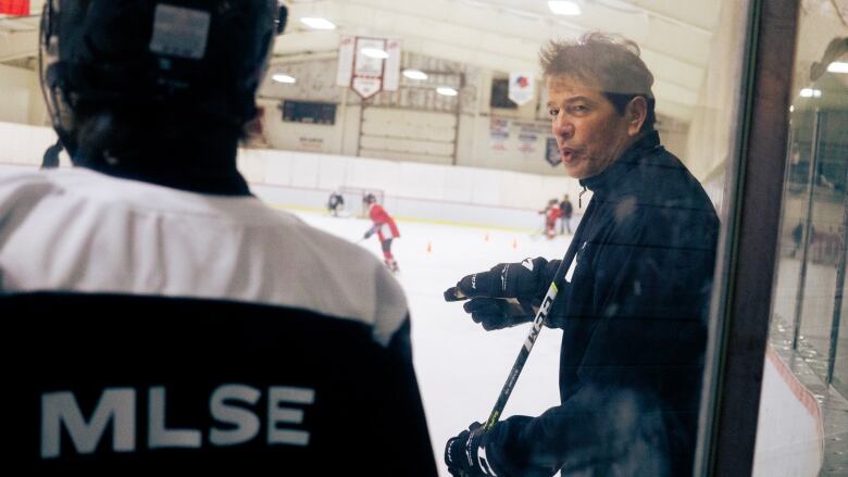 A photo taken through the glass of a hockey rink. In the foreground is a player with his back up against the boards. In focus is Ted Nolan, wearing all black.