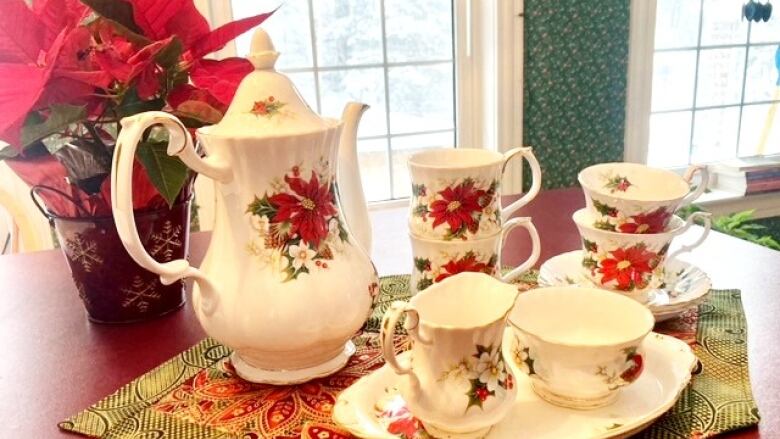 A coffee pop next to four tea cups, a plat, a creamer pot and a sugar dish. All of the pieces have poinsettias on them and are next to a poinsettia plant.