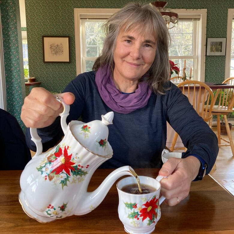 A woman pouring tea into a tea cup from a coffee pot with poinsettias on them.