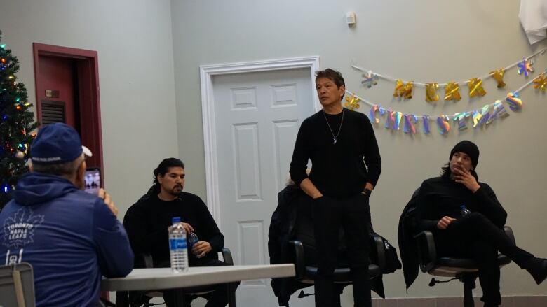 Two men sit and a third stands between them during a speech. 