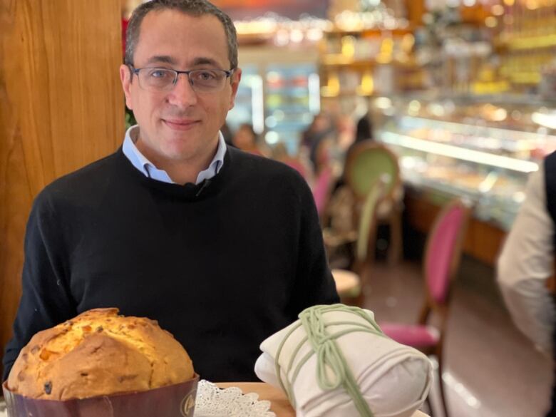 A smiling man wearing a sweater and glasses is shown at a table with a baked sweet bread and a white cloth pouch tied with a green string.