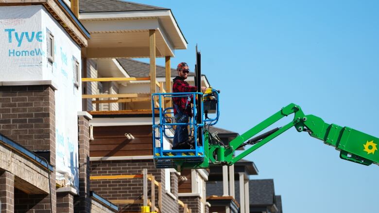 A man in a red and black jacket and a hard hat stands atop a cherry picker near a house under construction, with a blue sky in the background.