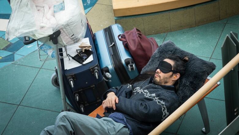 A bearded man sleeps next to a pile of luggage with a black eye mask covering his eyes and bright orange earplugs in his ears.