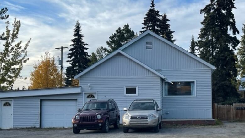 A blue house with a large, gravel driveway. There are two SUVs parked outside the property. There is blue sky and tall trees in the background.