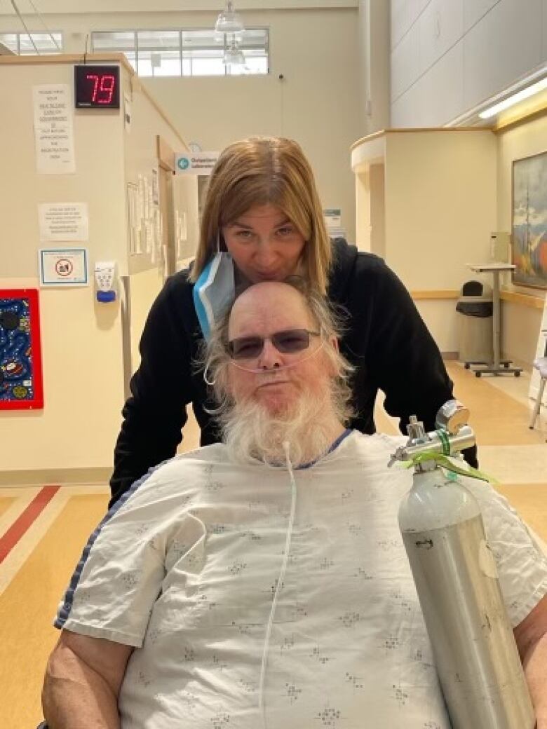 A man sits in a wheelchair wearing a hospital gown. He has white hair and sunglasses, and there are tubes connected to his nose. Behind him stands a woman resting her chin on the top of his head. The background shows they're in a hospital hallway.