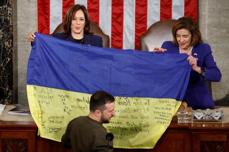 Individual in brown sweatshirt stands before a podium where two women hold a blue and yellow flag. 