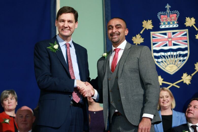 A tall white man and a South Asian man shake hands while being flanked by other people and the emblem of British Columbia.