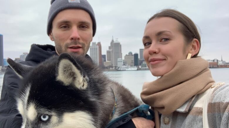 Husband and wife stand at Windsor's waterfront with a blue-eyes huskey. 