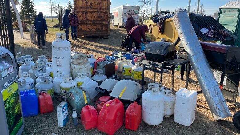 A group of propane tanks, jerry cans, and a barbeque sit outside in front of a dumpster and a large truck. A few people are milling about in the background. 