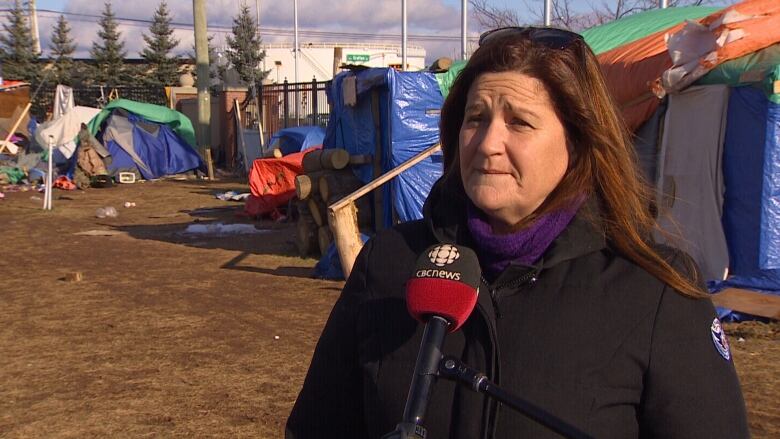 A woman doing an interview with CBC stands outside in front of a group of tents covered in tarps. 