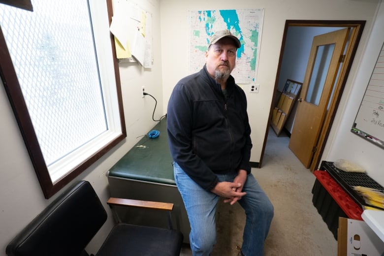 A man wearing blue jeans, a black zip-up sweater and a baseball cap sits on the edge of an office desk with his hands in his lap.