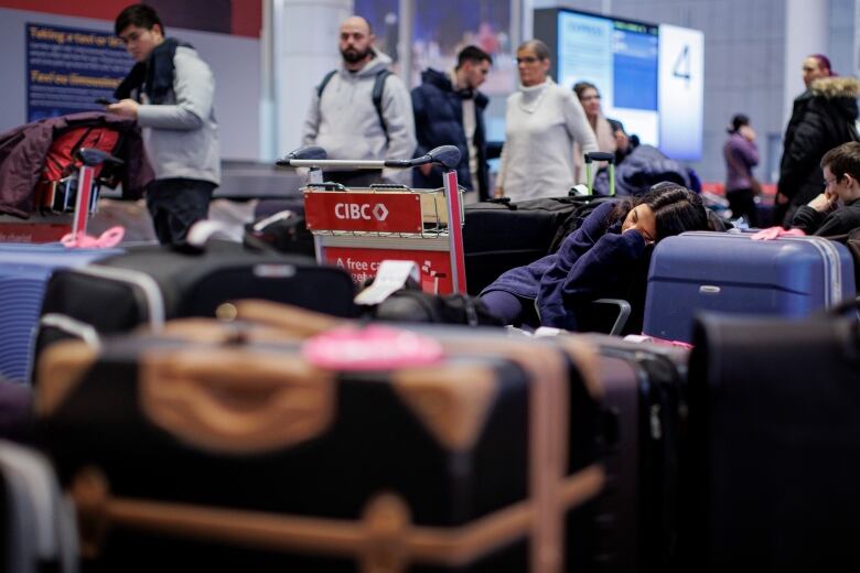 A woman is shown in a busy airport with luggage around her. She rests her head on her luggage.