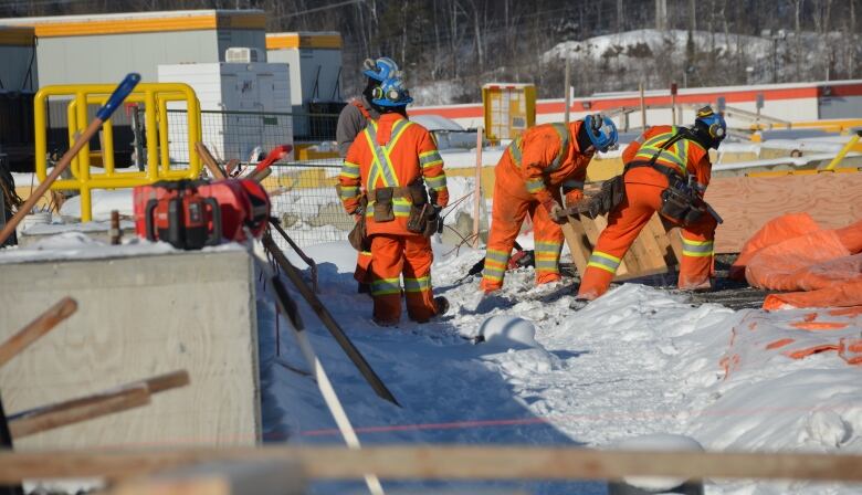 Construction workers in orange jackets and blue hard hats push a piece of wood into place, surrounded by a snowy landscape