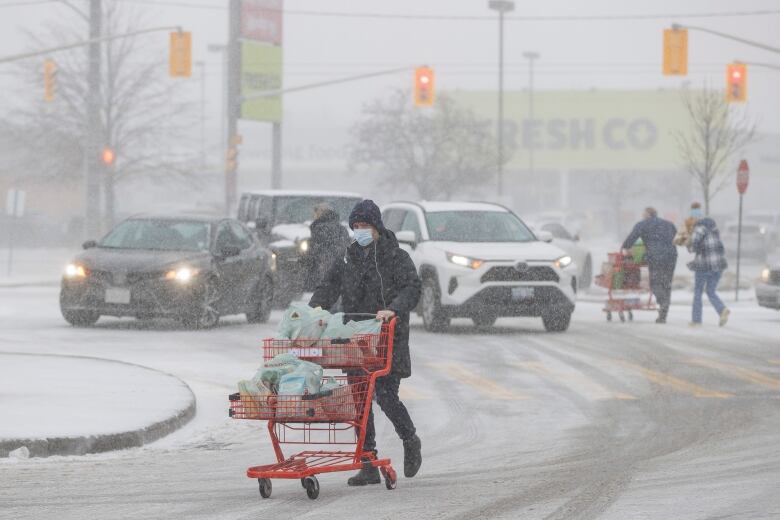 A person pushes a grocery cart full of grocery bags in a snowy parking lot of a grocery store.