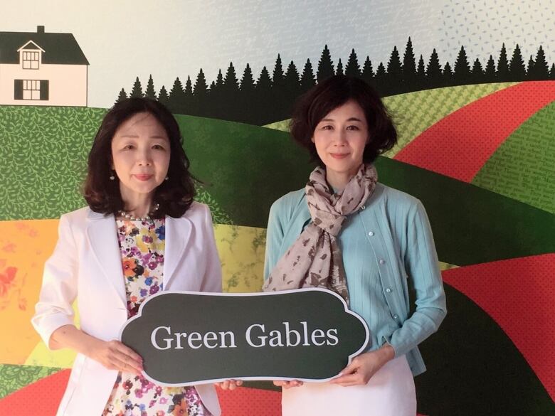 Two women stand in front of a painting, holding a sign that reads Green Gables. 