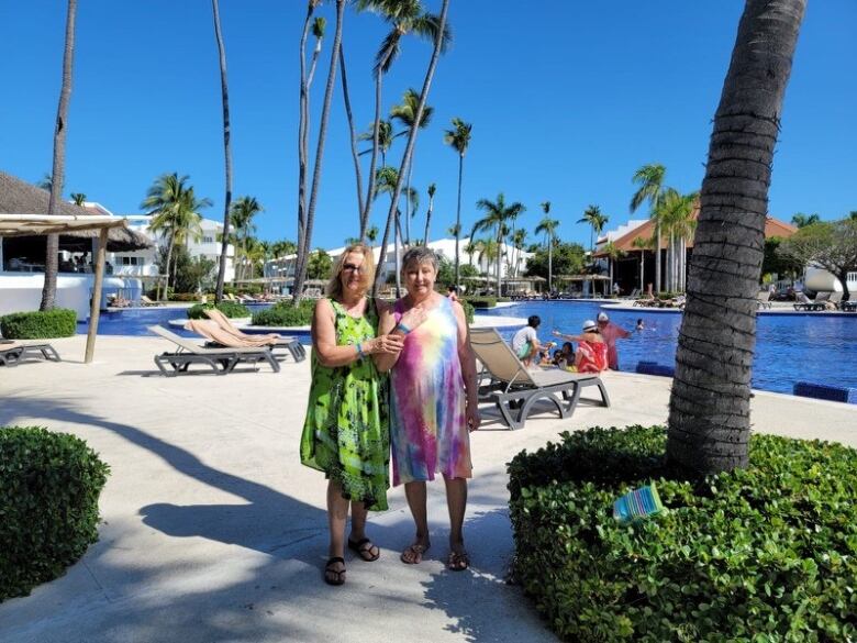 Two women stand, wearing sundresses, in front a pool. There are palm trees in the background. 