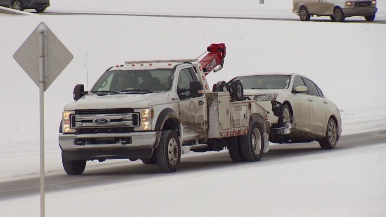 A white tow truck pulling a damaged white sedan on a snowy road.