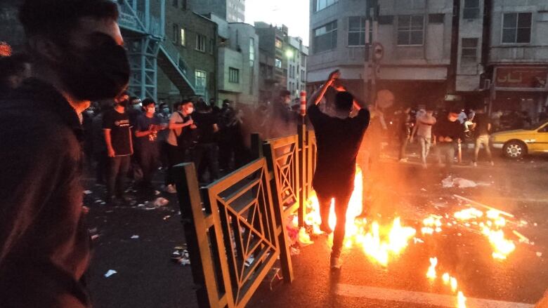 A person waves their arms in front of a fire in the street during a protest as other masked protesters look on. 