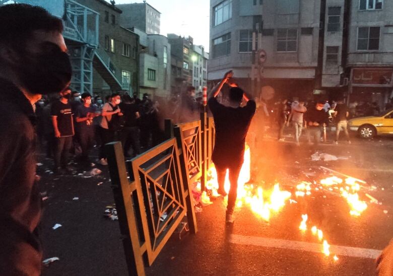 A person waves their arms in front of a fire in the street during a protest as other masked protesters look on. 