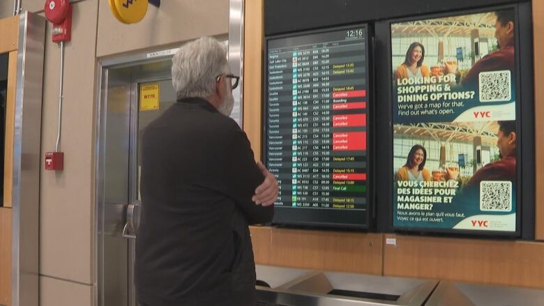 A man standing in front of an arrivals and departures board schedule at the Calgary International Airport.