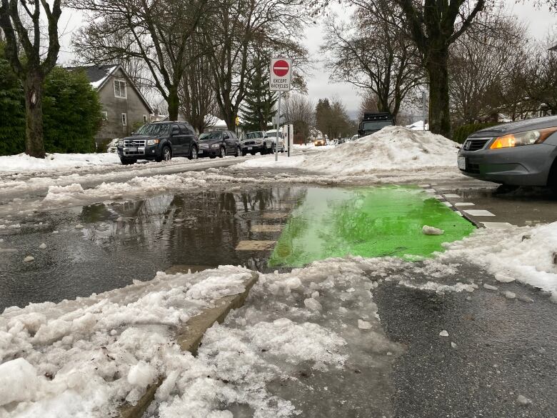 A pedestrian crossing has significant water pooling on a residential street, as cars are parked on the side of the road and slush is visible all around.