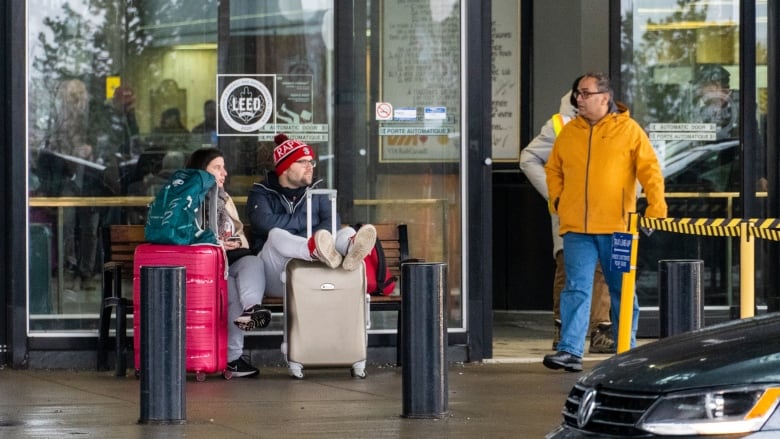 Two people sit on a bench with their feet up on their luggage while another person walks near them.