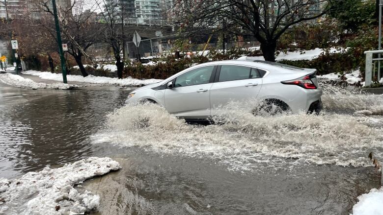 A car spews up water as it traverses a sludgy water-pooled road, with apartment buildings visible in the background.