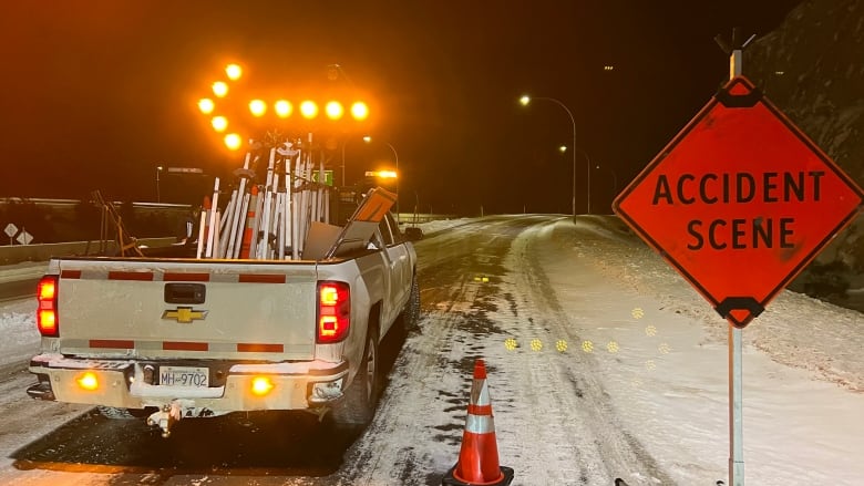 A pickup truck with a flashing leftward arrow parked on a snowy highway. A sign next to it reads 'ACCIDENT SCENE'. Orange traffic cones are visible.