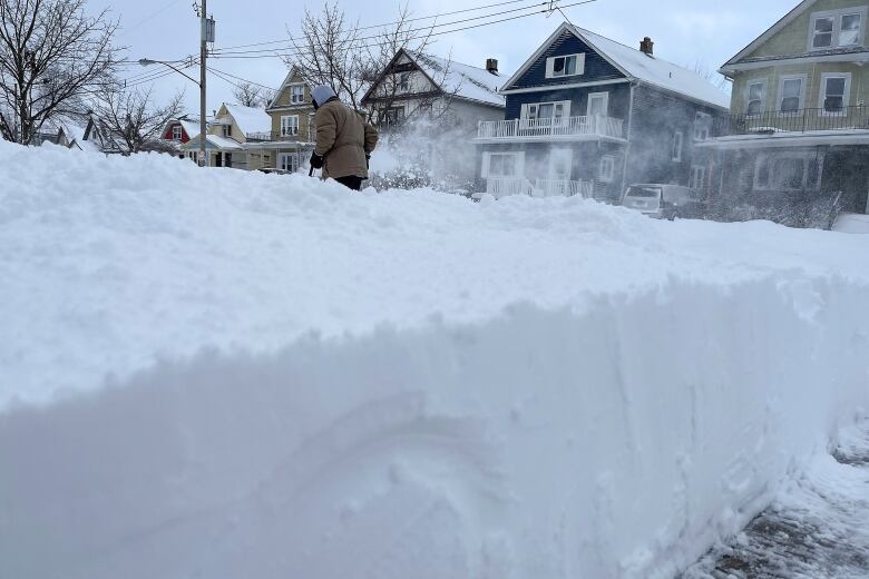 A man pushes a snow blower behind a high snowbank next to a driveway. 